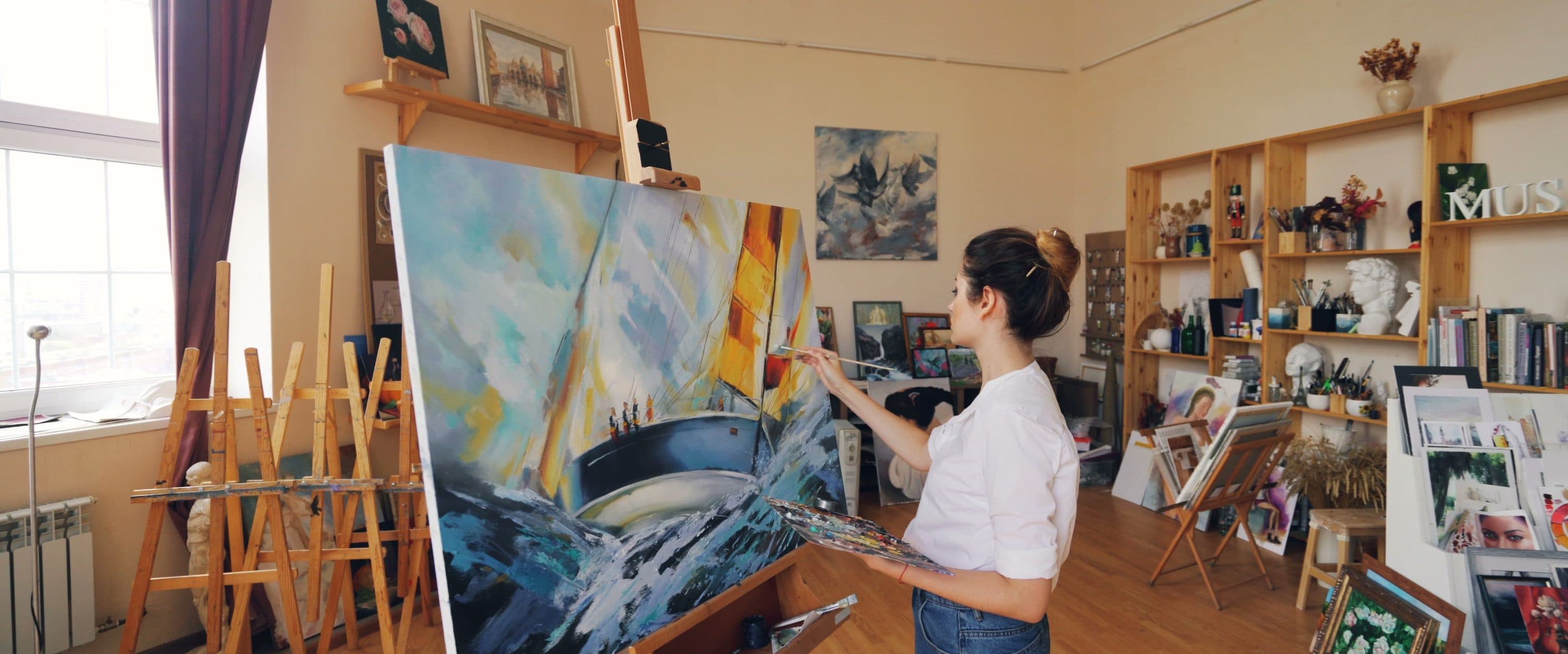 An artist painting a seascape in a sunlit studio lined with shelves of work.